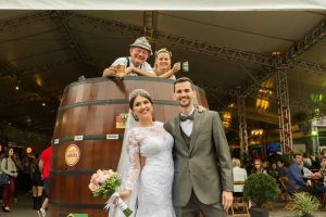 Paula e Rafael posam ao lado de um barril enquanto visitantes da Schützenfest aparecem ao fundo