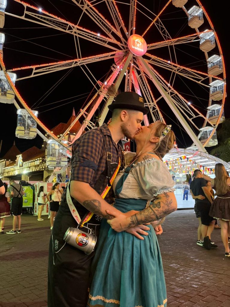 Casal vestido com trajes alemães se beija em frente à roda-gigante iluminada