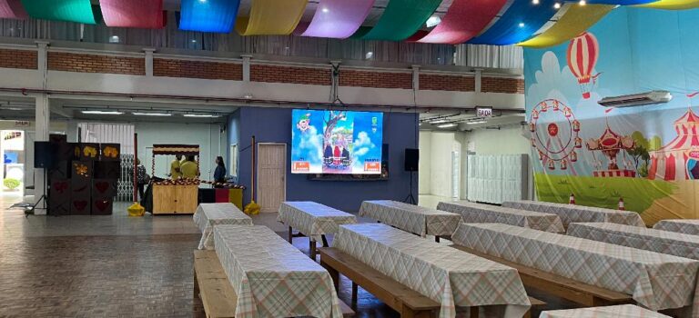 Telões instalados no Parque de Eventos exibem conteúdo da Schützenfest 2025 em espaço decorado com mesas e bandeiras coloridas